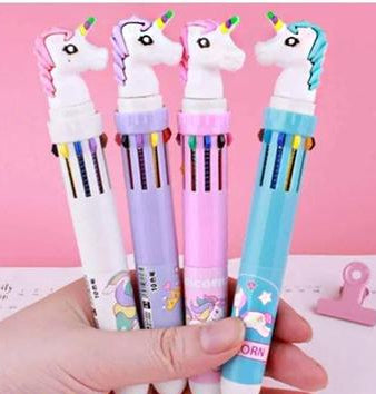 Four unicorn-shaped multi-color pens held in a hand against a pink background. The pens have white, lavender, pink, and blue bodies with colorful accents and cartoon unicorn designs.