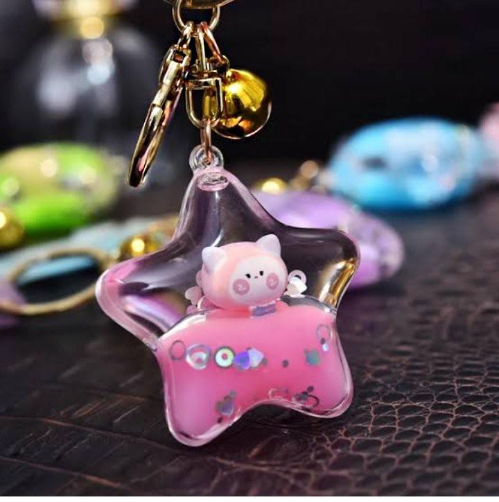 A star-shaped acrylic keychain charm with a pink cat figure inside a pink liquid-filled center, attached to gold-tone hardware and a small gold bell, photographed on a dark textured surface with blurred colorful keychains in the background.