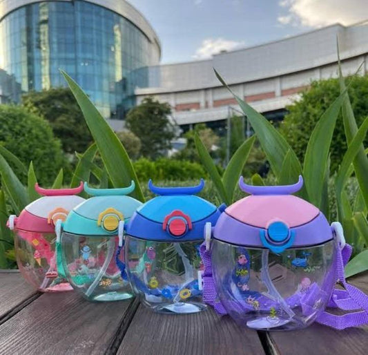 Four reusable water bottles with colorful lids and purple shoulder straps, lined up on a wooden table with green plants and a modern glass building in the background.