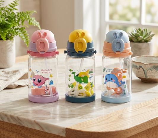 Three transparent baby water bottles with colorful lids and cartoon animal prints, arranged on a light wood countertop in a bright indoor setting with plants and a window in the background.